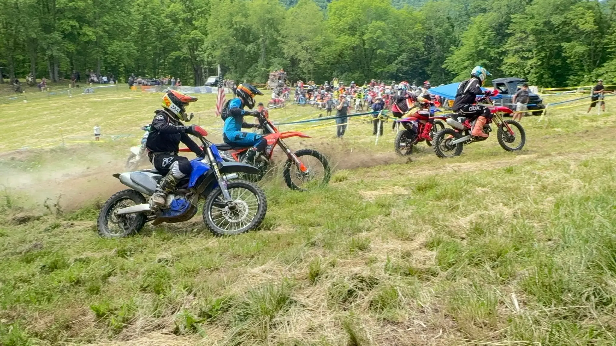 Riders racing across a grassy hillside past a long line of spectators and pop-up tents