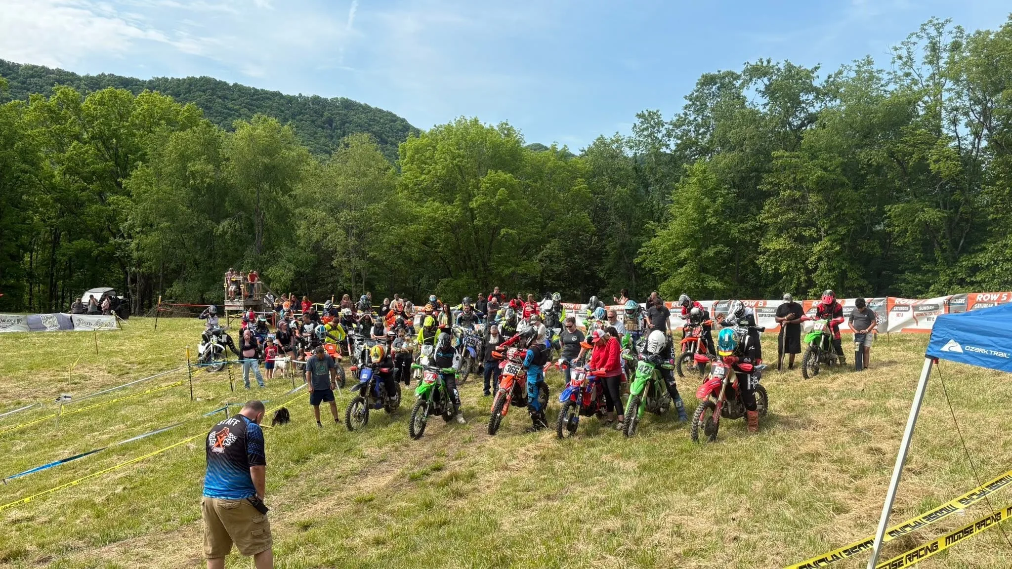 A big crowd of riders and bikes gathered for the start on a hillside, with wooded mountains behind
