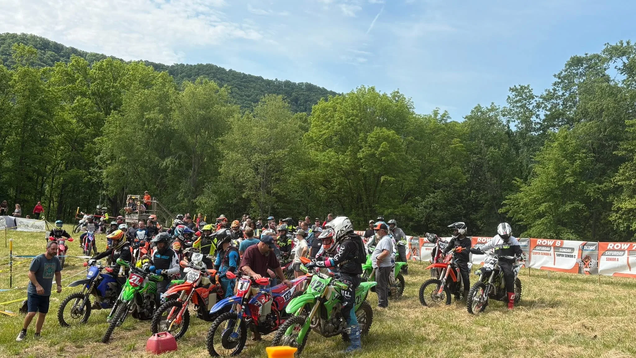 Riders and bikes packed together at the start line on a grassy field, with wooded mountains behind