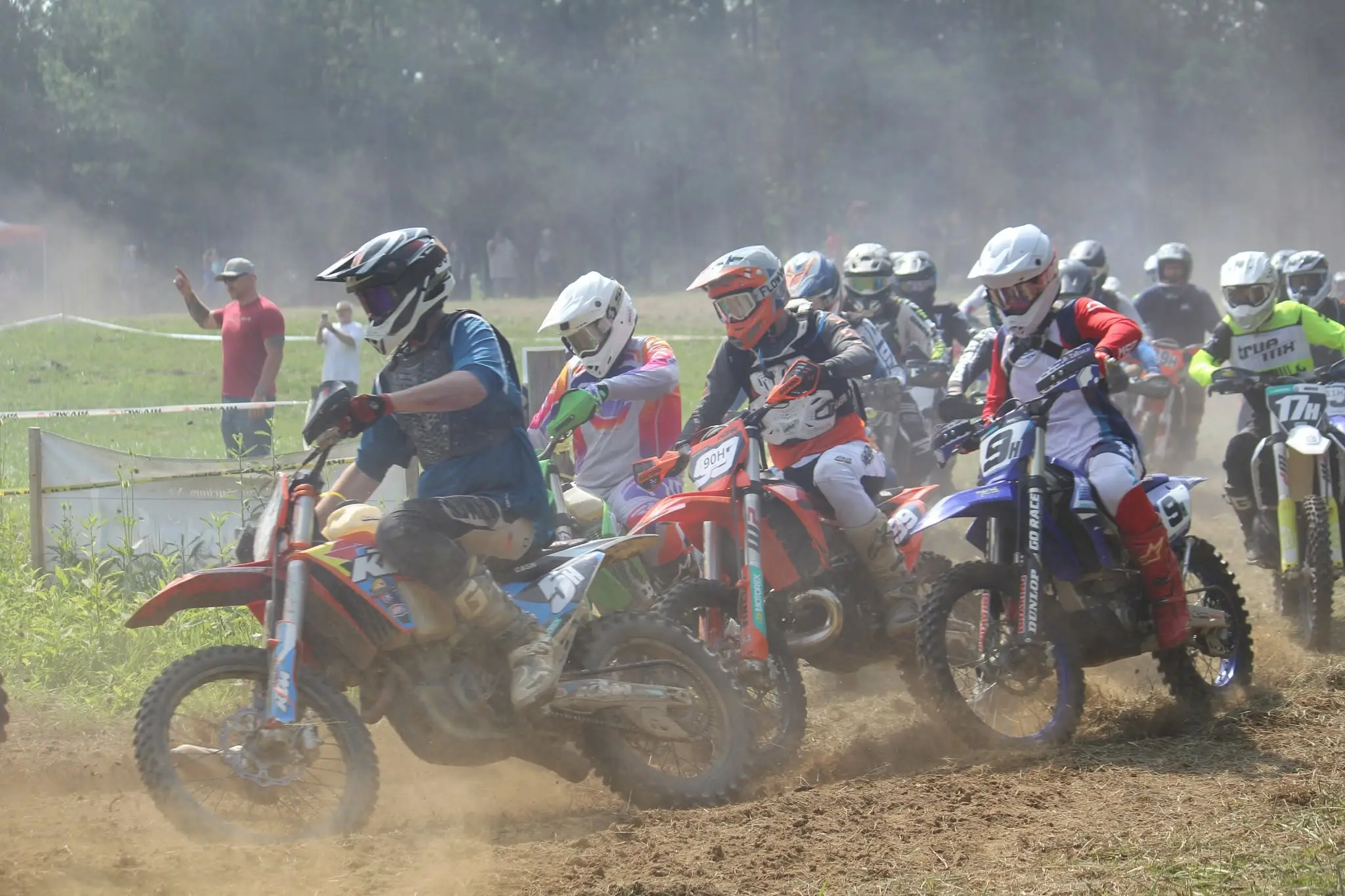 A pack of riders charging off the start in a cloud of dust, with spectators watching from behind the tape