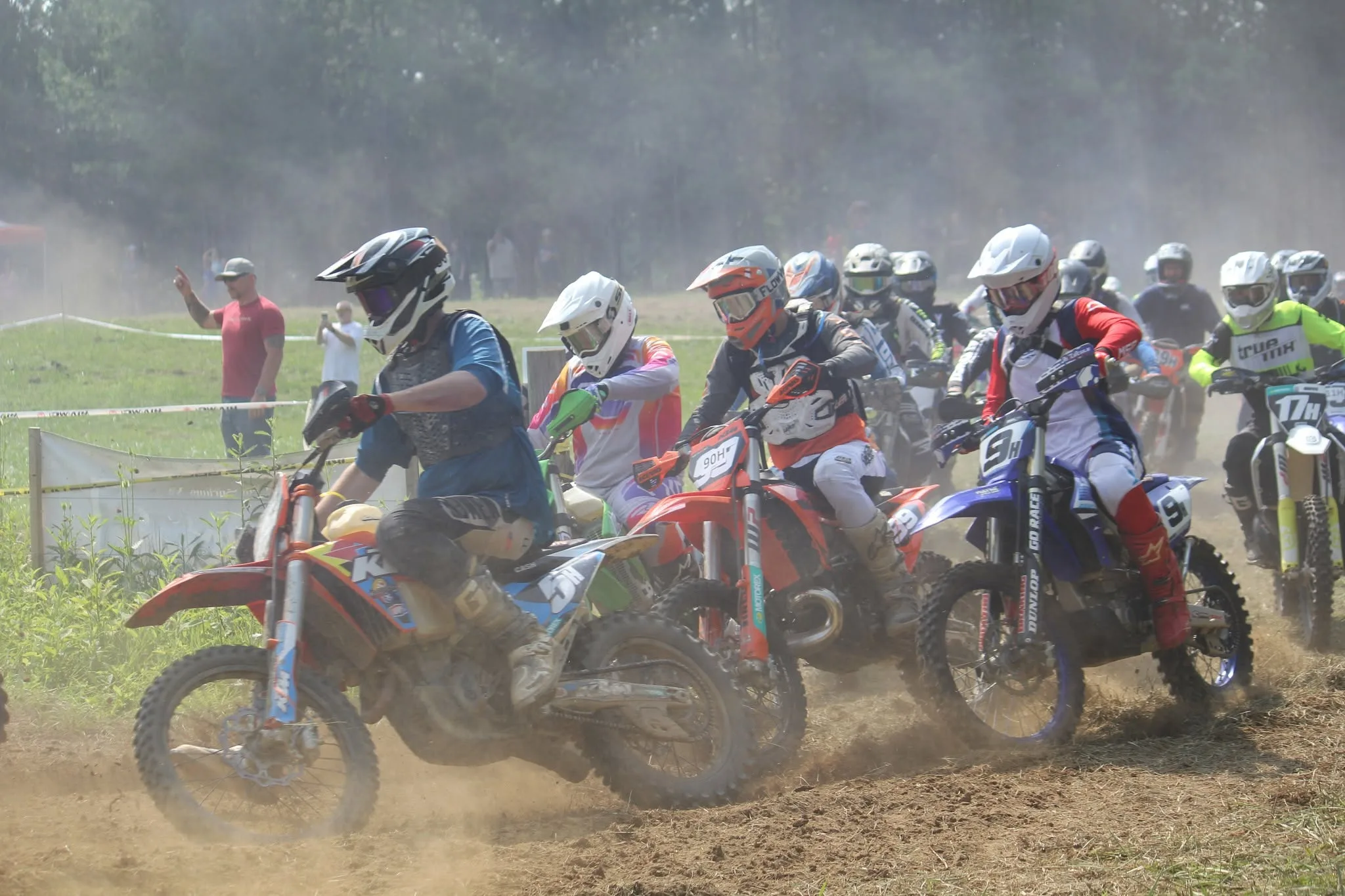 A pack of riders charging off the start in a cloud of dust, with spectators watching from behind the tape
