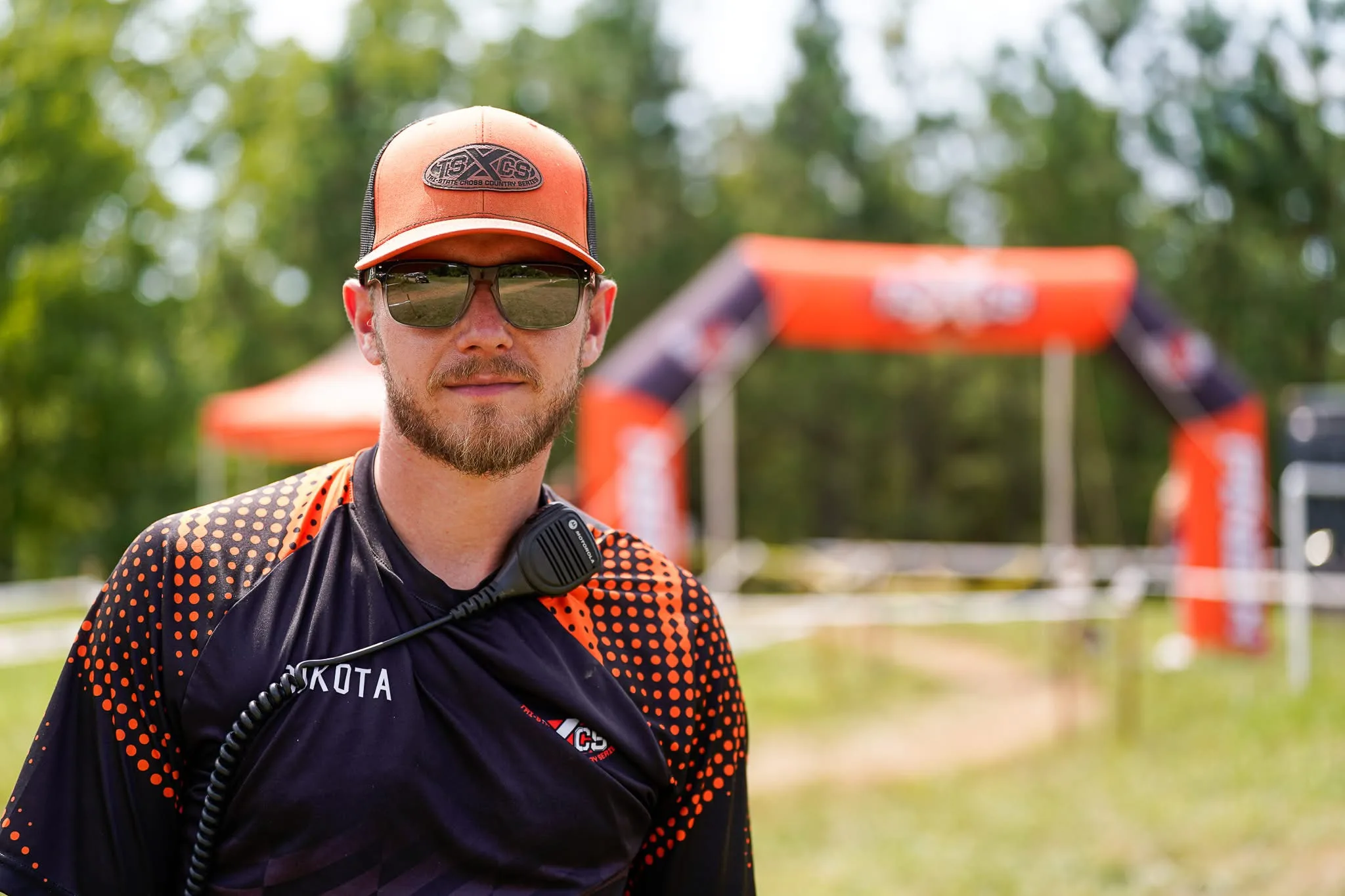 Dakota Weddle in a TSXCS cap and jersey with a radio on his shoulder, the orange TSXCS start arch behind him